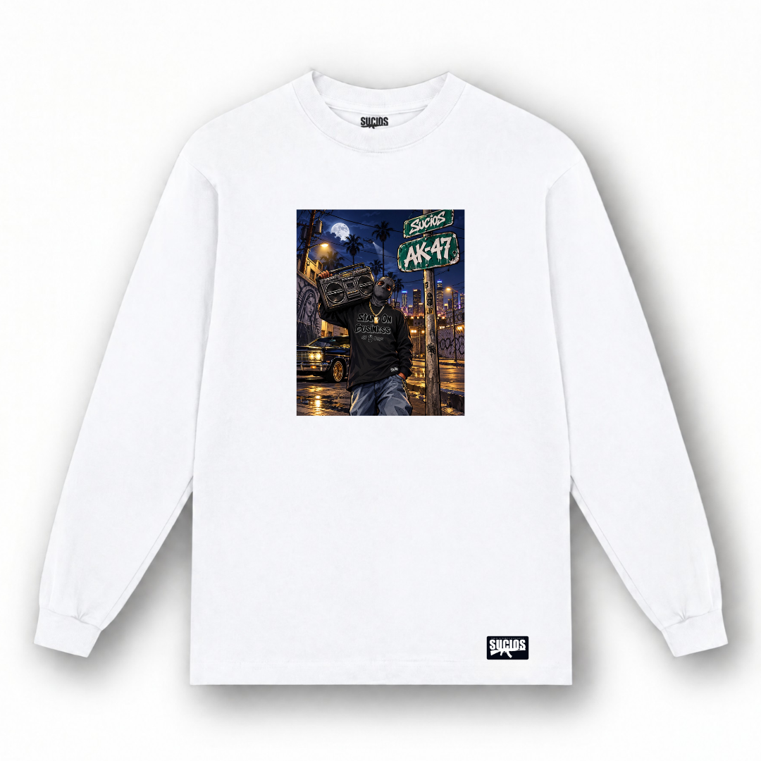 A white long-sleeve crewneck t-shirt with a central graphic print of a person carrying a boombox on their shoulder in a city street at night, featuring a green 'AK-47' sign. The brand 'SUCIOS' is visible on the inner neck label and as a small black logo patch near the bottom hem.